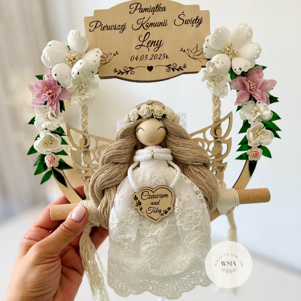 Personalizowana pamiątka Pierwszej Komunii Świętej w formie anioła z makramy na drewnianej obręczy. Rękodzieło ozdobione białymi i różowymi kwiatami, idealne jako prezent komunijny dla dziewczynki od rodziców chrzestnych lub dziadków.