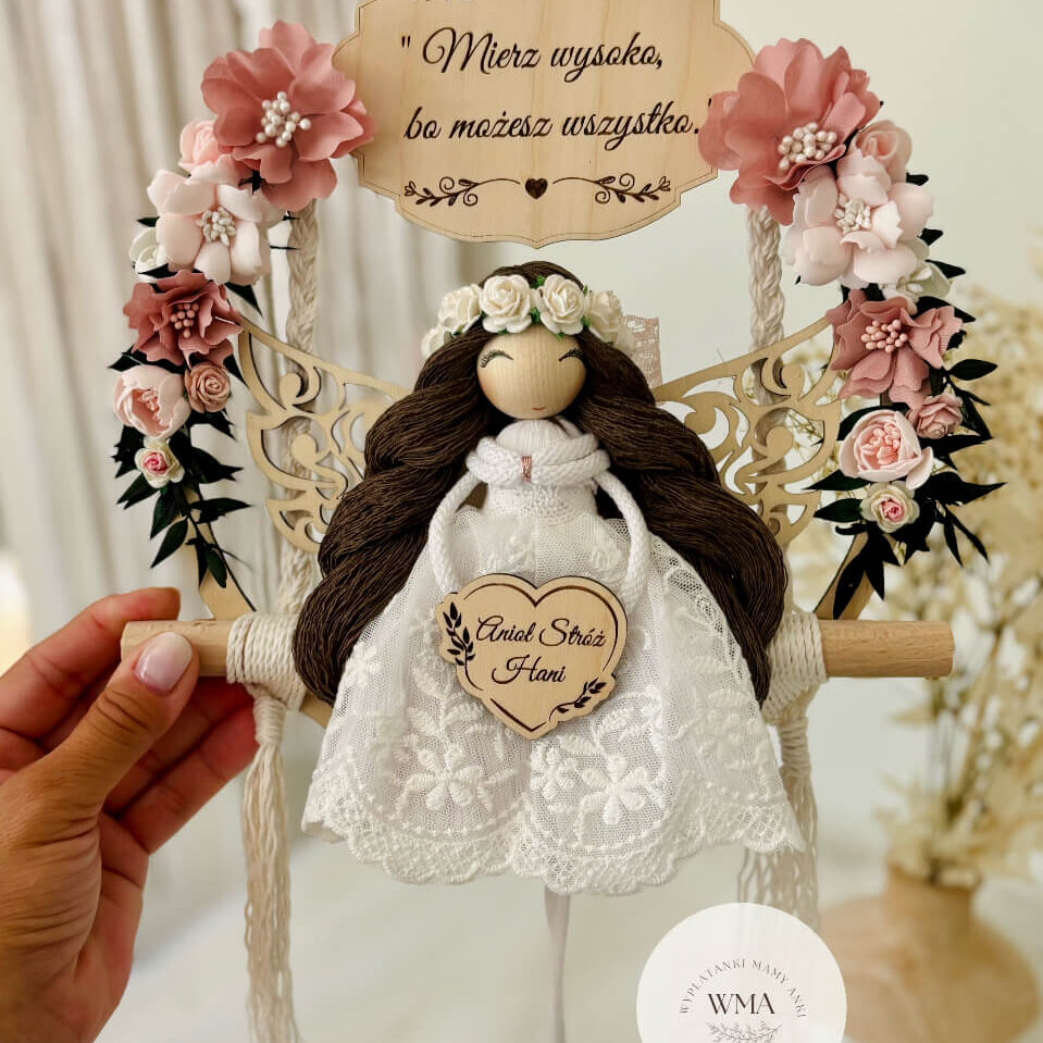 Ręcznie wykonany anioł stróż z makramy z ciemnymi włosami w białej koronkowej sukience. Personalizowana pamiątka na drewnianej obręczy z cytatem motywacyjnym "Mierz wysoko, bo możesz wszystko". Idealny prezent dla dziewczynki w stylu boho.