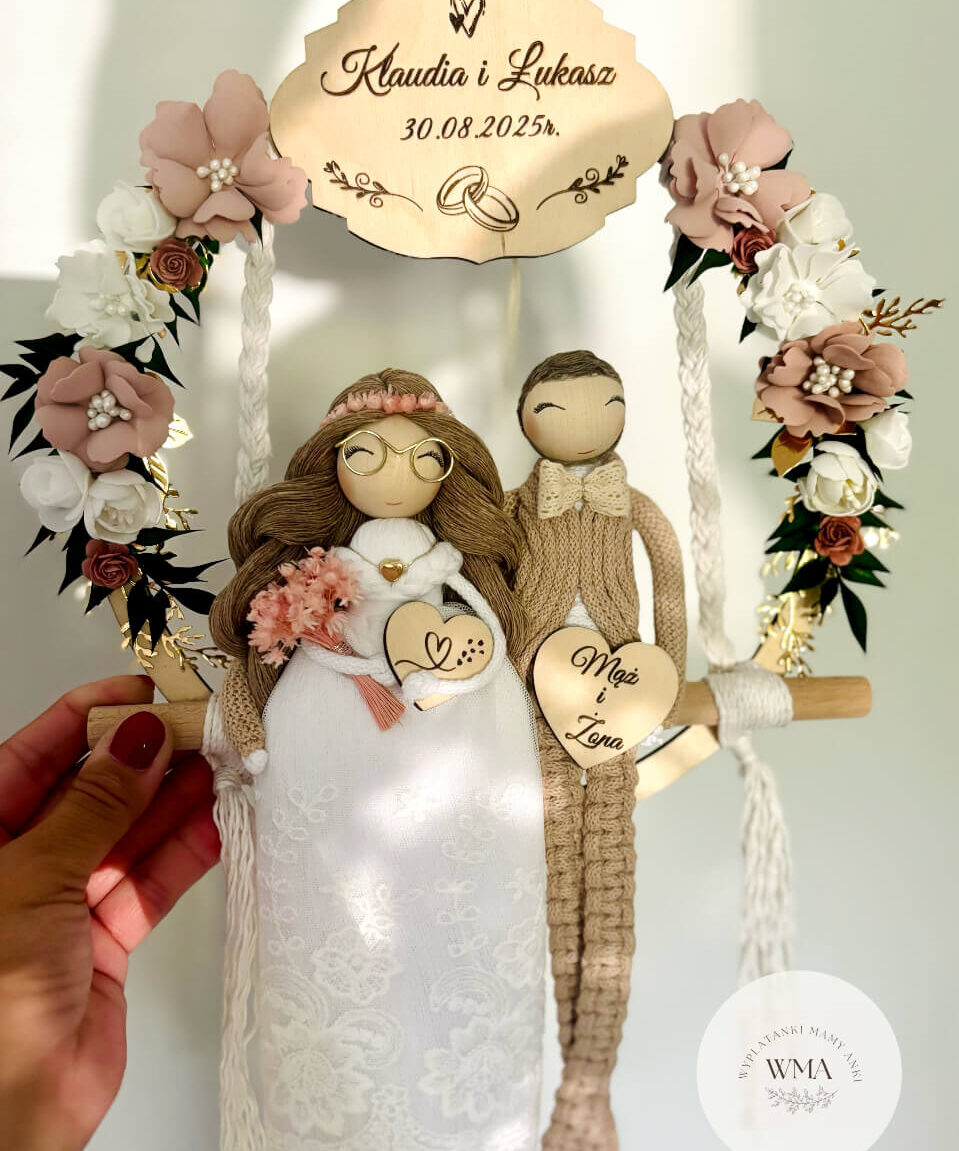 Personalizowana dekoracja ślubna na huśtawce makramowej z figurkami pary młodej - panna młoda w okularach z brązowymi włosami w różowym wianku i koronkowej sukni z różowym bukietem, pan młody w beżowym stroju z makramy z kokardką, złote lustrzane tabliczki z imionami i datą ślubu oraz sercem z napisem Mąż i Żona, dekoracja z różowych białych i bordowych kwiatów ze złotymi akcentami, handmade