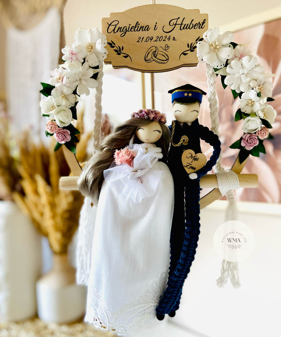 Personalizowana dekoracja ślubna na huśtawce makramowej z figurkami pary młodej - panna młoda z brązowymi włosami w fioletowym wianku i sukni koronkowej z różowym bukietem, pan młody w granatowym mundurze policjanta z czapką i złotymi akcentami z makramy, drewniana tabliczka z imionami i datą ślubu, serce z napisem Mąż i Żona, dekoracja z białych i różowych kwiatów, handmade