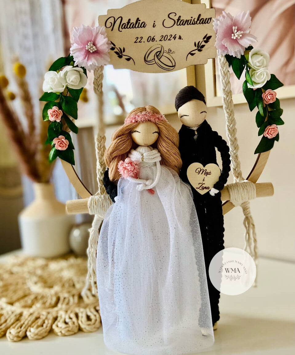Personalizowana dekoracja ślubna na huśtawce makramowej z figurkami pary młodej - panna młoda z brązowymi włosami w różowym wianku i tiulowej sukni brokatowej z różowym bukietem, pan młody w czarnym stroju z makramy, drewniana tabliczka z imionami i datą ślubu, serce z napisem Mąż i Żona, dekoracja z różowych i białych kwiatów z zielenią, handmade