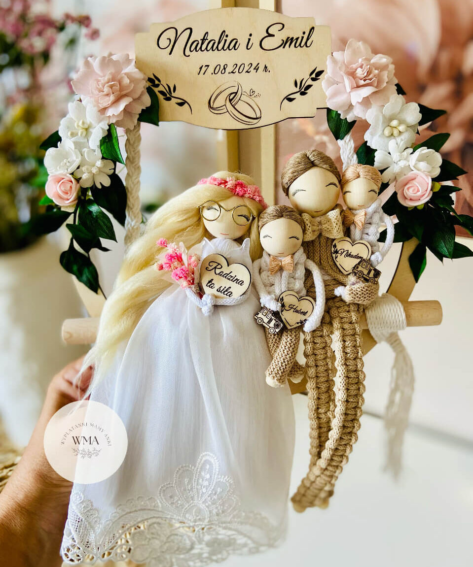 Personalizowana dekoracja ślubna na huśtawce makramowej z figurkami całej rodziny - panna młoda w okularach z długimi blond włosami w różowym wianku i sukni koronkowej, pan młody w beżowym stroju z makramy, dwoje dzieci z drewnianymi sercami z imionami i miniaturami pojazdów, tabliczka z napisem Rodzina to siła, drewniana tabliczka z imionami i datą ślubu, dekoracja z różowych i białych kwiatów, handmad