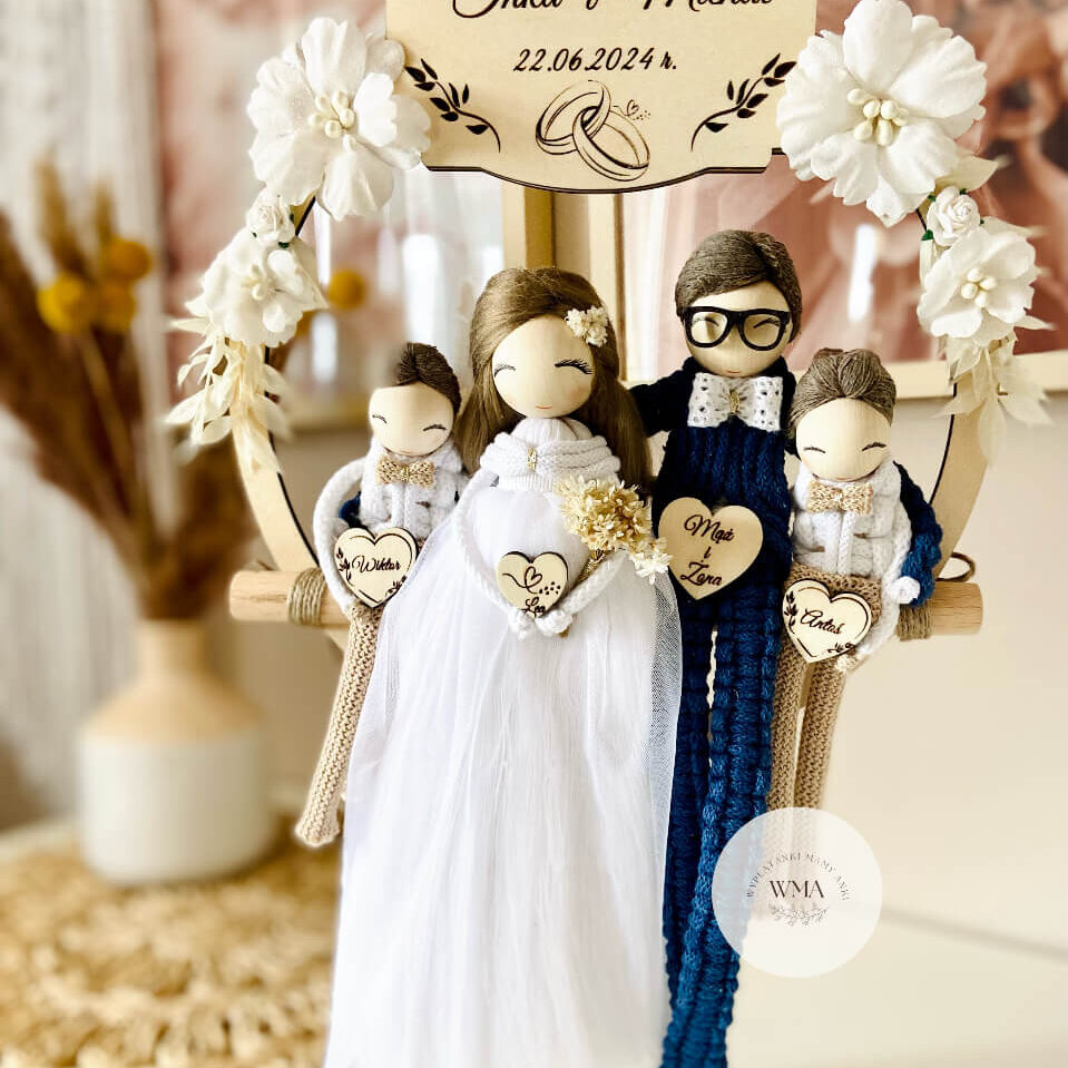 Personalizowana dekoracja ślubna na huśtawce makramowej z figurkami całej rodziny - panna młoda w tiulowej sukni z kwiatkiem we włosach, pan młody w okularach w granatowym stroju z makramy, dwoje dzieci z drewnianymi sercami z imionami, drewniana tabliczka z imionami rodziców i datą ślubu, dekoracja z białych kwiatów, handmade