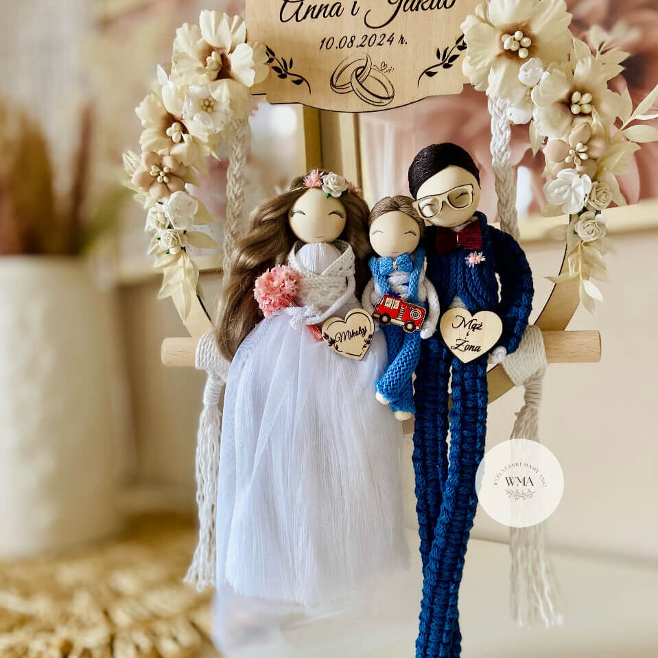 Personalizowana dekoracja ślubna na huśtawce makramowej z figurkami rodziny - panna młoda z brązowymi włosami w tiulowej sukni z różowym bukietem, pan młody w okularach w granatowym stroju z makramy z bordową muszką, figurka dziecka w niebieskim stroju z drewnianym wozem strażackim, drewniana tabliczka z imionami i datą ślubu, serce z napisem Mąż i Żona, dekoracja z kremowych kwiatów, handmade