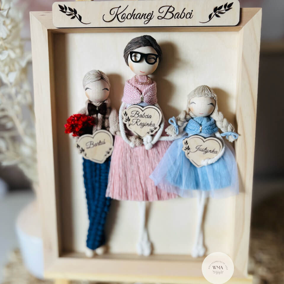 Personalizowana ramka w sosnowej ramie z napisem Kochanej Babci z trzema figurkami z makramy - babcia w okularach w różowej sukience z frędzli z wnukiem w granatowym stroju z czerwonym bukietem i wnuczką w błękitnej tiulowej sukience z warkoczem, drewniane serca z imionami, wyjątkowy prezent dla babci na urodziny lub Dzień Babci, handmade