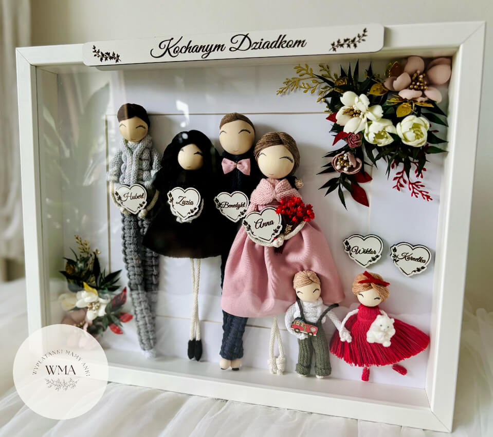 Personalizowana biała ramka z napisem Kochanym Dziadkom z sześcioma figurkami całej rodziny z makramy - cztery dorosłe figurki w szarym czarnym granatowym i różowym stroju oraz dwoje małych dzieci z drewnianymi sercami z imionami i miniaturą wozu strażackiego, dekoracja z białych czerwonych i złotych kwiatów, wyjątkowy prezent dla dziadków, handmade