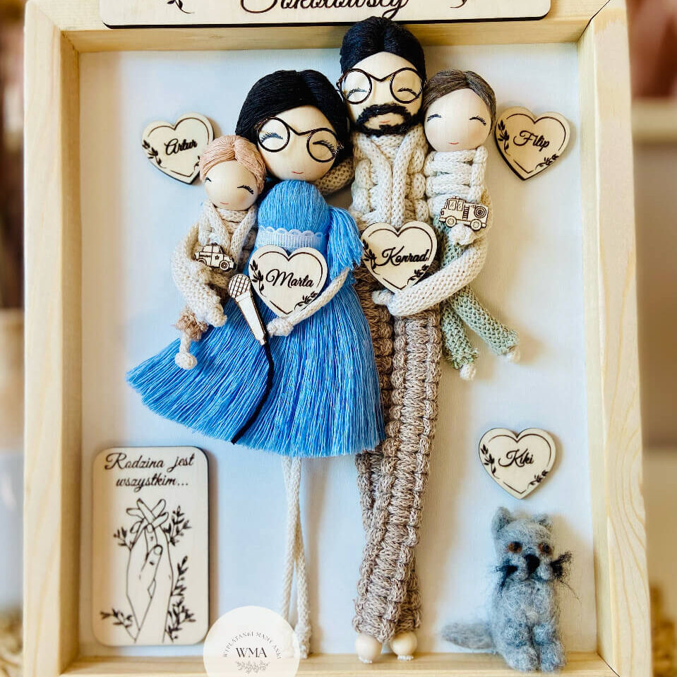 Personalizowana ramka rodzinna w sosnowej ramie z nazwiskiem rodziny z czterema figurkami z makramy i filcowym kotem - mama w okularach w niebieskiej sukience z frędzli trzymająca niemowlę, tata z brodą w okularach trzymający drugie dziecko, filcowy szary kot z imieniem, drewniane serca z imionami i tabliczka Rodzina jest wszystkim, handmade