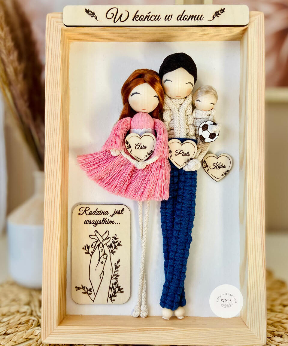 Personalizowana ramka w sosnowej ramie z napisem W końcu w domu z trzema figurkami rodziny z makramy - mama w różowej sukience z frędzli z rudymi włosami, tata w granatowym stroju trzymający niemowlę z piłką nożną, drewniane serca z imionami i tabliczka z napisem Rodzina jest wszystkim, wyjątkowy prezent na przeprowadzkę lub narodziny dziecka, handmade