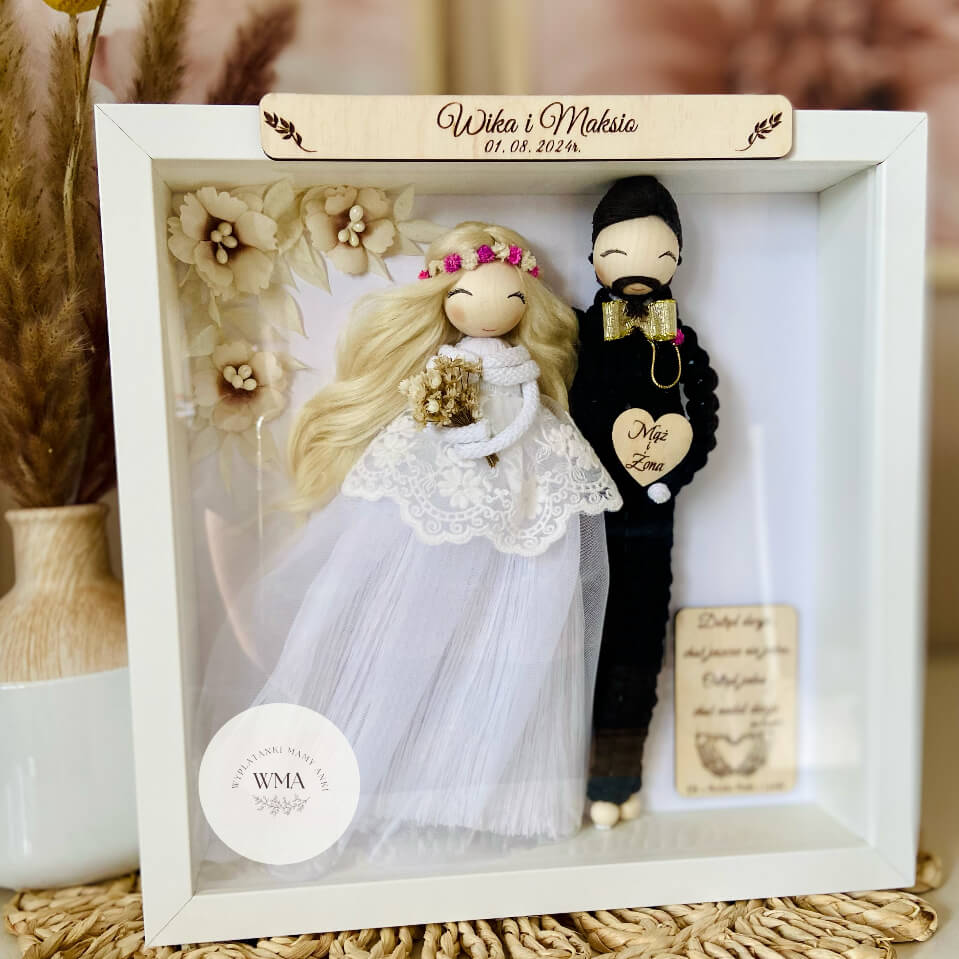 Personalizowana ramka ślubna z figurkami pary młodej - panna młoda z długimi blond włosami w różowym wianku i tiulowej sukni z koronką, pan młody z brodą w czarnym stroju z makramy ze złotą muszką, drewniana tabliczka z imionami i datą ślubu, serce z napisem Mąż i Żona, dekoracja z beżowych kwiatów, handmade