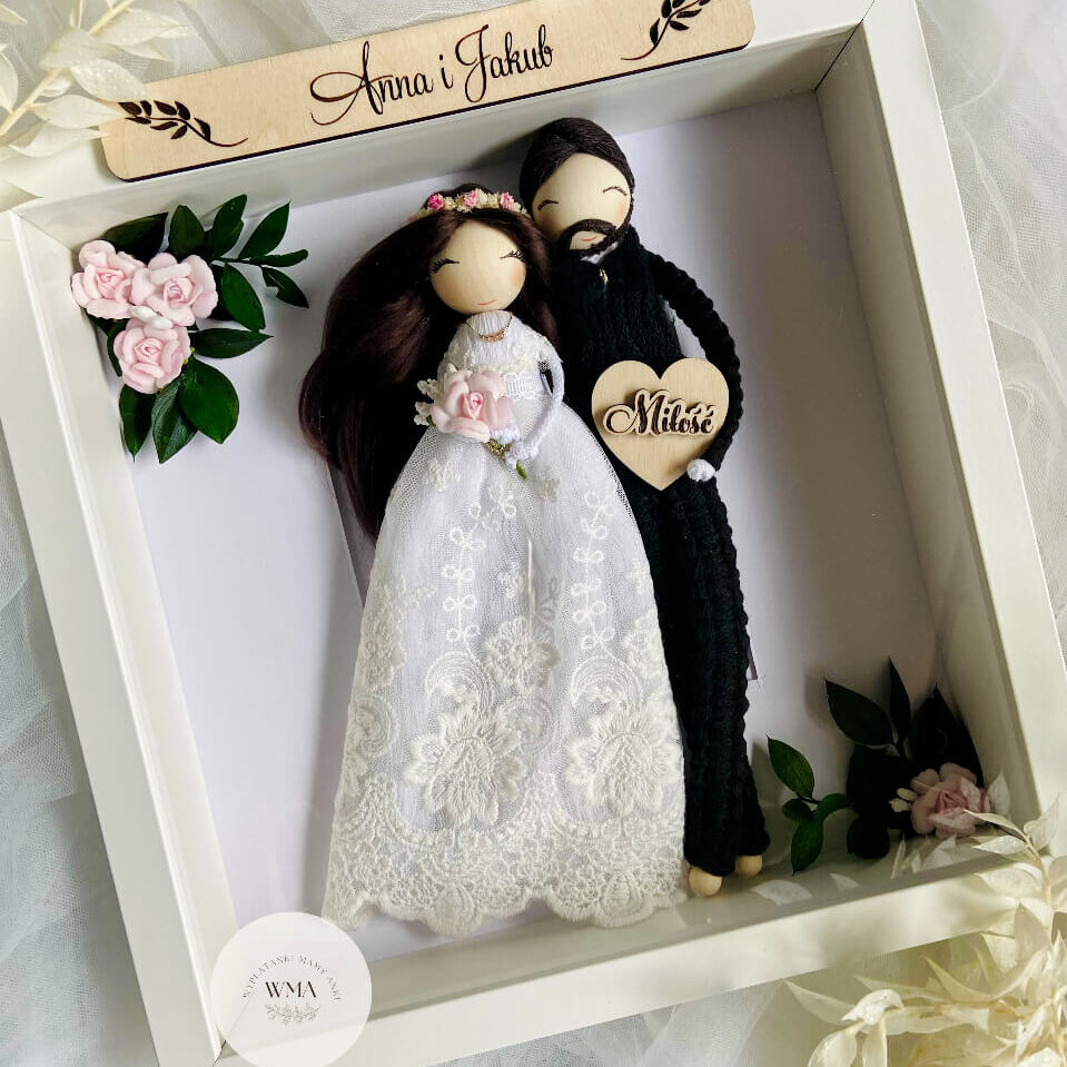 Personalizowana ramka ślubna z figurkami pary młodej - panna młoda z ciemnymi włosami w różowym wianku i sukni koronkowej z różowym bukietem, pan młody z brodą w czarnym stroju z makramy, drewniana tabliczka z imionami, serce z napisem Miłość, dekoracja z różowych róż i zielonych gałązek, handmade