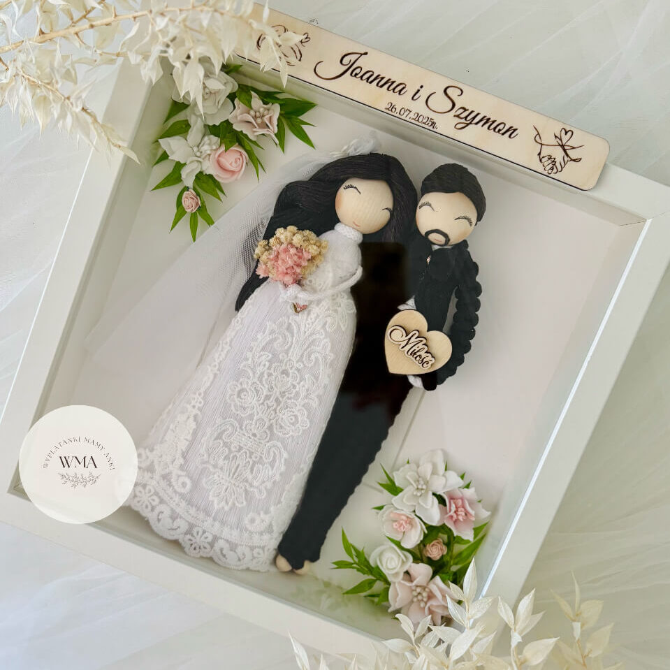 Personalizowana ramka ślubna z figurkami pary młodej - panna młoda z czarnymi włosami w welonie i sukni koronkowej z różowym bukietem, pan młody z brodą w czarnym stroju z makramy, drewniana tabliczka z imionami i datą ślubu, dekoracja z różowych i białych kwiatów z zielenią, handmade