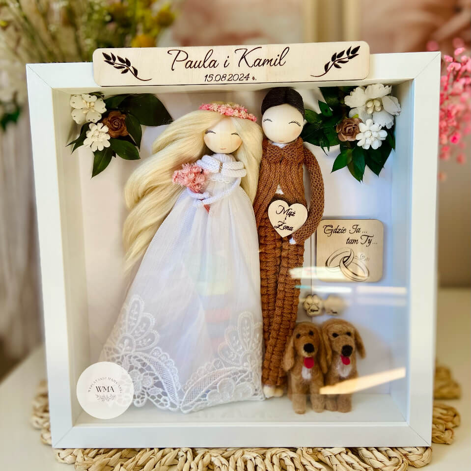Personalizowana ramka ślubna z figurkami pary młodej i dwiema miniaturami psów - panna młoda z długimi blond włosami w różowym wianku i tiulowej sukni koronkowej, pan młody w brązowym stroju z makramy, dwa filcowe psy, drewniana tabliczka z imionami i datą ślubu, serce z napisem Mąż i Żona, dekoracja z białych kwiatów i zieleni, handmade