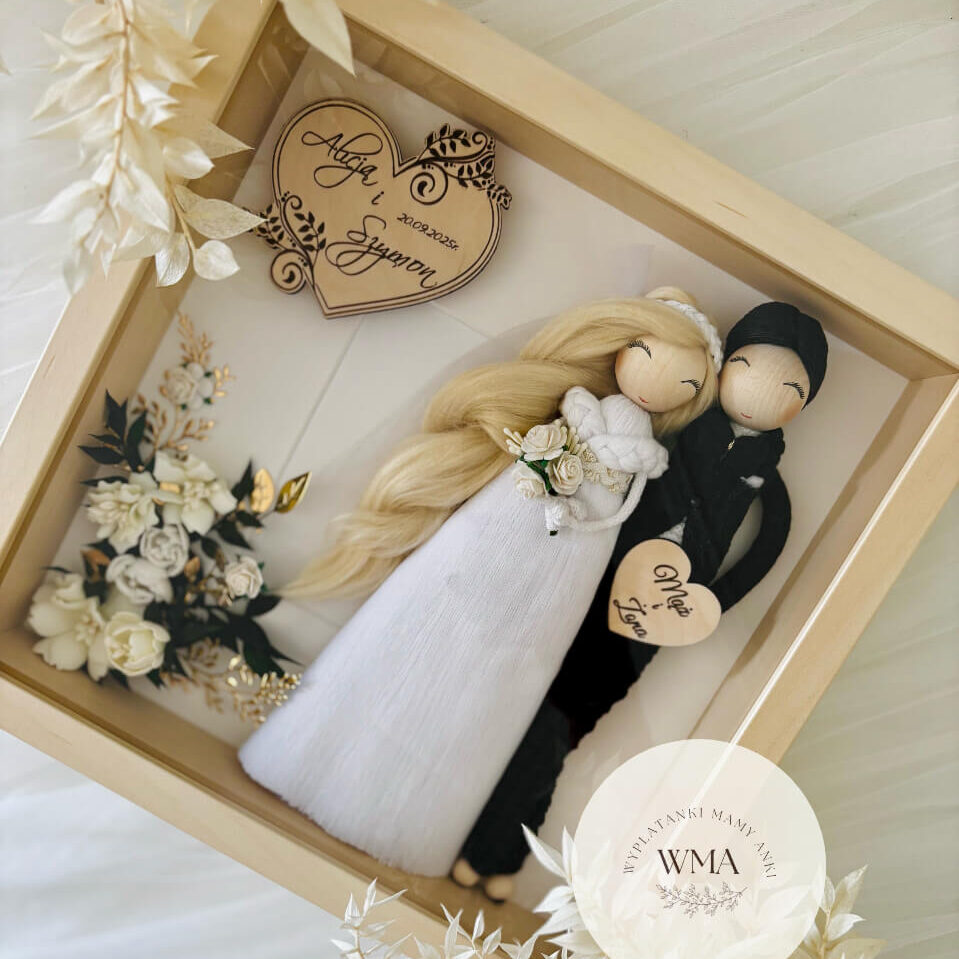 Personalizowana ramka ślubna w naturalnej drewnianej ramie z figurkami pary młodej - panna młoda z długimi blond włosami w białej tiulowej sukni z białymi różami, pan młody w czarnym stroju z makramy, drewniane serce z imionami i datą ślubu, serce z napisem Ona i On, dekoracja z białych kwiatów i złotych akcentów, handmade