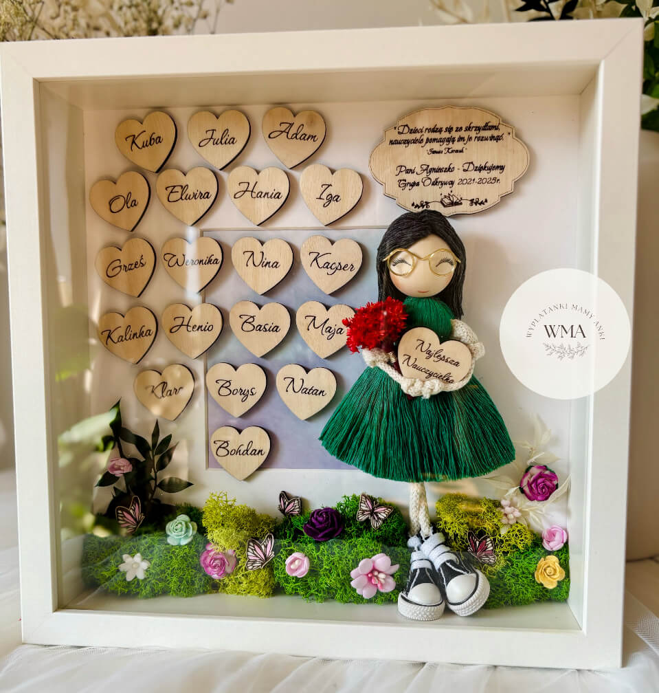 Personalizowana drewniana ramka z podziękowaniem dla nauczyciela z ręcznie robianą laleczką z czarnymi włosami w zielonej sukience z frędzlami i złotymi okularami trzymającą serduszko Najlepsza Nauczycielka, drewniane serduszka z imionami dzieci, dekoracja z mchu i kolorowych kwiatów z motylami, prezent od klasy