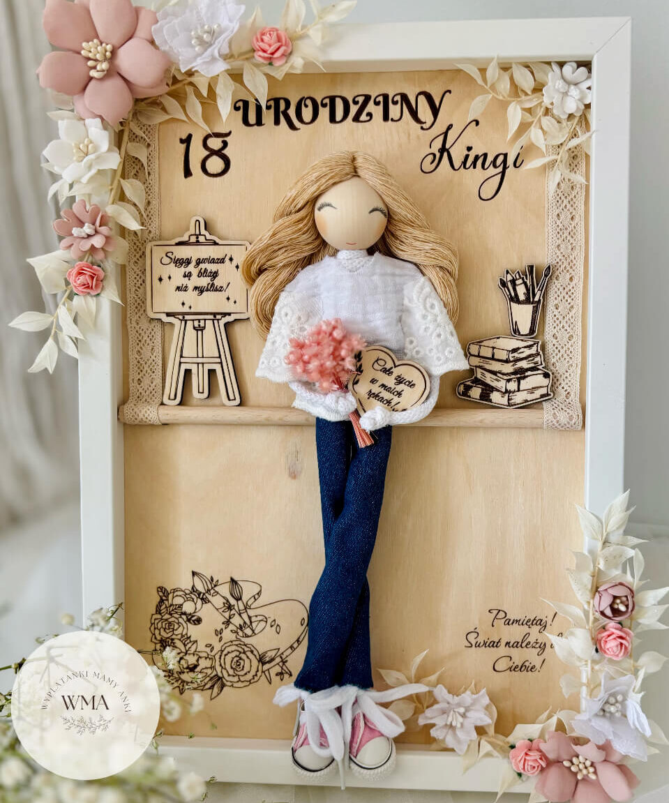 Personalizowana drewniana ramka na osiemnastkę z ręcznie robioną laleczką w dżinsach i białym sweterku trzymającą różowy bukiet, drewniane elementy ze sztalugą i książkami, biało-różowe kwiaty na ramie, elegancki prezent urodzinowy